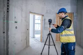 A person wearing safety equipment adjusts a laser leveling tool on a tripod in a concrete room.