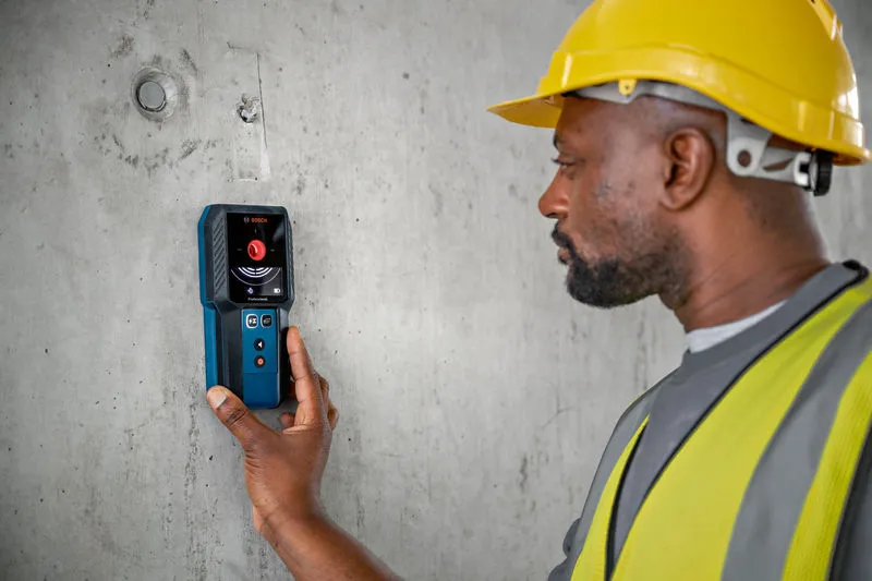 A person wearing safety equipment uses a detector to scan a concrete wall.