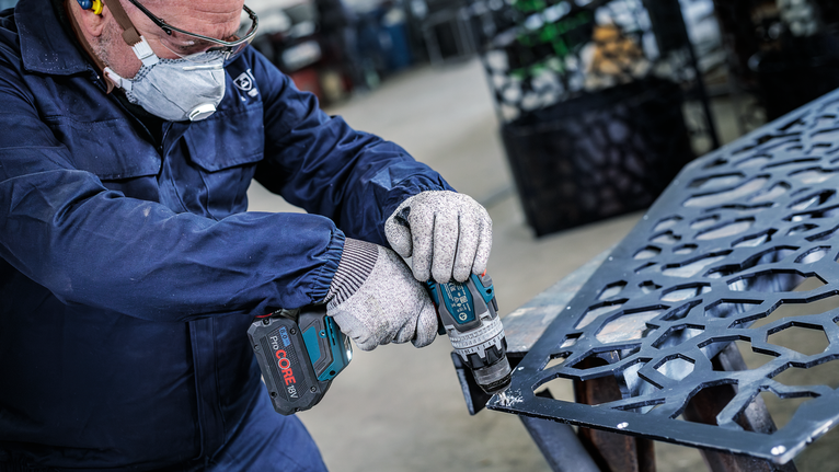 A person wearing safety equipment drills into a patterned metal sheet.