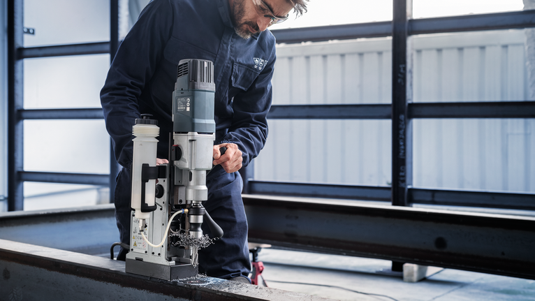 A person wearing safety equipment operates a magnetic drill on a steel beam.