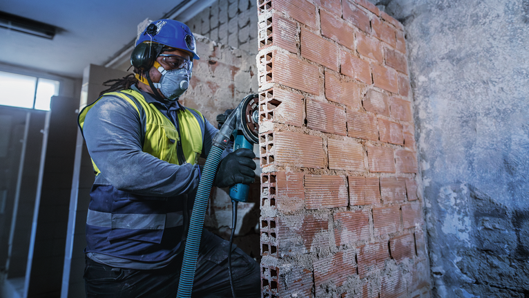 A person wearing safety equipment cuts into a brick wall using a power tool.