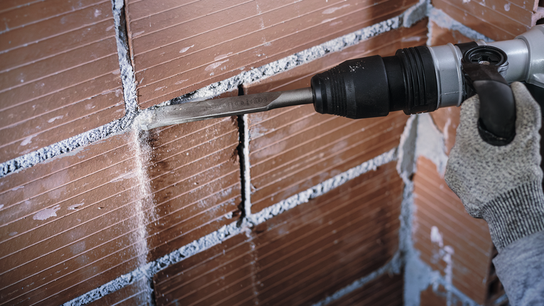 Person wearing safety equipment uses a power chisel to remove mortar from a brick wall.
