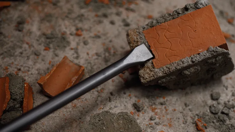 Chisel breaking a red brick block on a concrete surface.