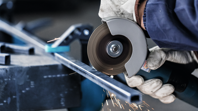 A worker wearing safety equipment cuts a metal bar with an angle grinder.