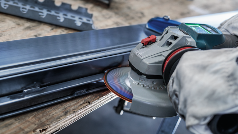 Person wearing safety equipment grinds a metal beam with an angle grinder.