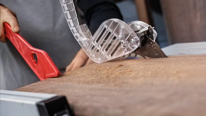 Person uses a push stick to guide wood through a table saw with safety guard.