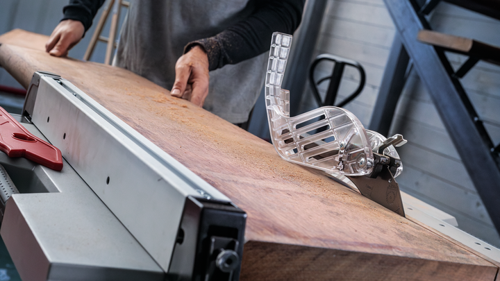 Person pushing a large wooden board through a table saw in a workshop.
