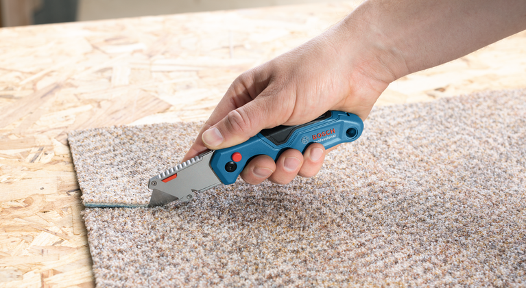 Person cuts carpet with a utility knife on a wooden surface.