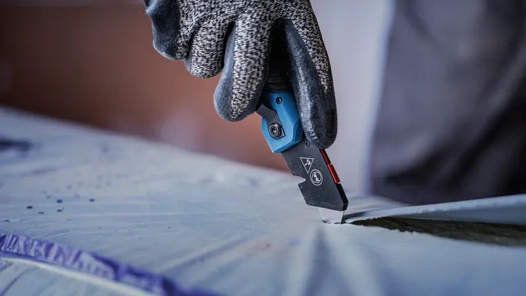 Person wearing safety equipment cuts plastic sheeting with a utility knife.
