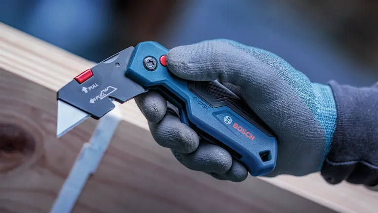 Person wearing safety equipment holds a utility knife near a wooden beam.