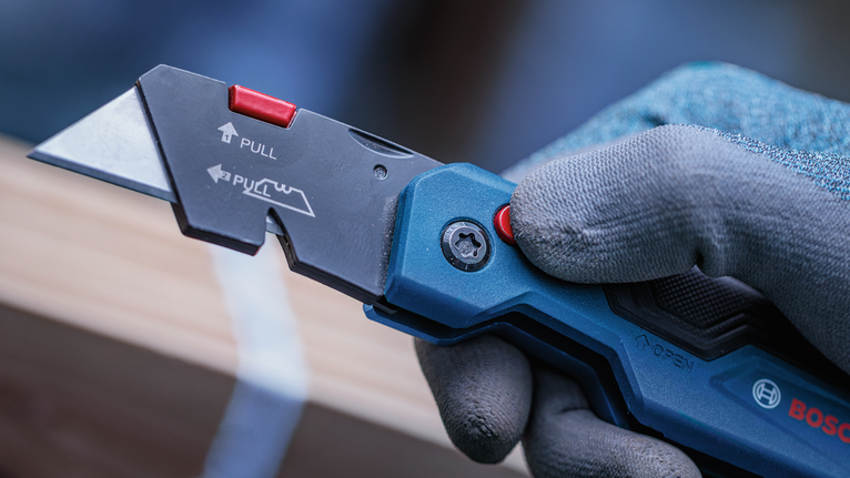 Person wearing safety equipment holding a utility knife near a wooden surface.