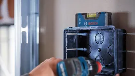 Person uses a power drill to install an electrical box under a leveling tool.