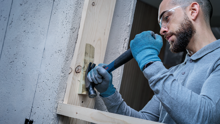 Person wearing safety equipment uses a hammer and chisel on a wooden plank.