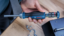 Person holding a multi-bit screwdriver near scattered screws on a wooden table.