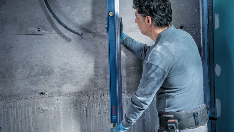 Person wearing safety equipment checks wall alignment with a long spirit level.