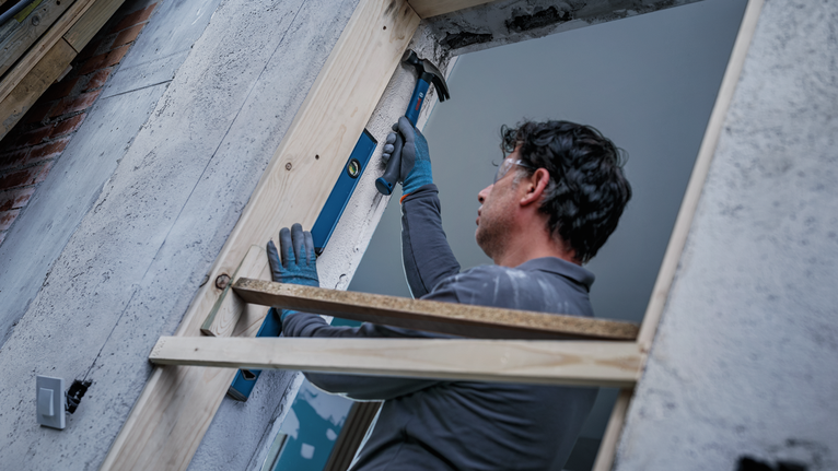 Person wearing safety equipment uses a drill to install a wooden frame in a wall opening.