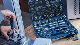 Person securing a metal component with a screwdriver, socket tool set open on the table.