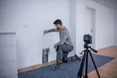 Personne agenouillée marquant un mur, guidée par un outil de nivellement laser sur un trépied.