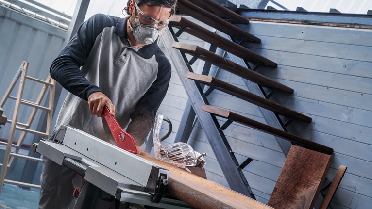 Une personne portant un équipement de sécurité utilise une scie à table pour couper du bois à l'intérieur.