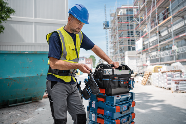 Une personne portant un équipement de sécurité range une sacoche à outils sur des boîtes à outils empilées sur un chantier de construction.