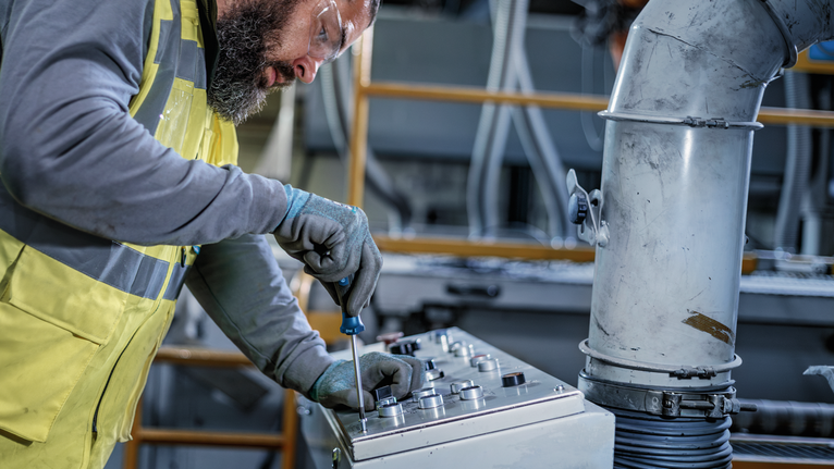 Une personne portant un équipement de sécurité utilise un tournevis sur un panneau de commande industriel.