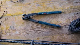 Une pince coupante en acier repose sur un établi en bois, à côté de fils enroulés et de barres d'armature.
