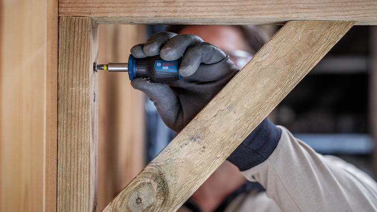Une personne portant un équipement de sécurité visse une vis dans un cadre en bois à l'aide d'un outil électrique.