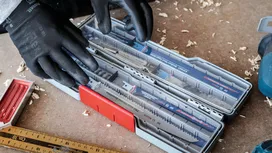 Una persona, que lleva puesto el equipo de seguridad, organiza las hojas de sierra en una caja de plástico sobre un banco de trabajo.