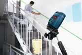 Una herramienta de nivelación láser proyecta una línea verde mientras una persona prepara una escalera para pintarla.