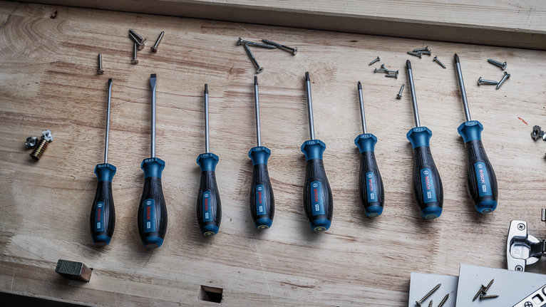 Ocho destornilladores con mangos azules dispuestos sobre un banco de trabajo de madera, cerca de tornillos dispersos.