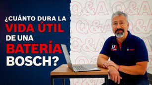 Hombre con barba sentado en un escritorio, con laptop, fondo azul y texto visible.