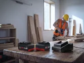Turvavarustust kandev inimene puurib töökojas puitu, laual elektritööriista akud.