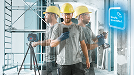 Mees kollase kiivri ja halli särgiga kasutab nutitelefoni ehitusplatsil.