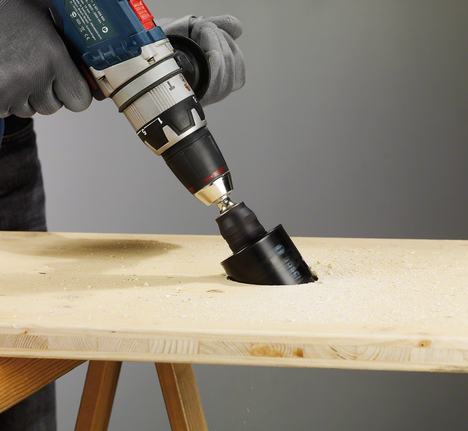Worker wearing safety equipment drills a large hole in wood with a hole saw.
