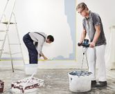 A person wearing safety equipment mixes paint with a stirring mechanism, while another paints a wall.