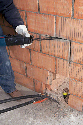 Person wearing safety equipment uses a reciprocating saw to cut a brick wall.