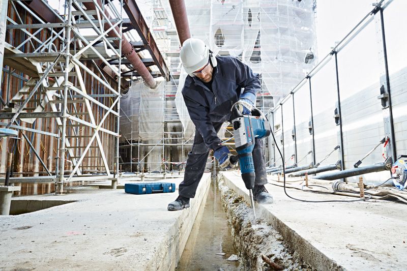 A person wearing safety equipment uses a rotary hammer to break concrete at a construction site.