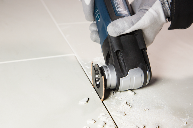 Person wearing safety equipment cuts a ceramic tile using an oscillating power tool.