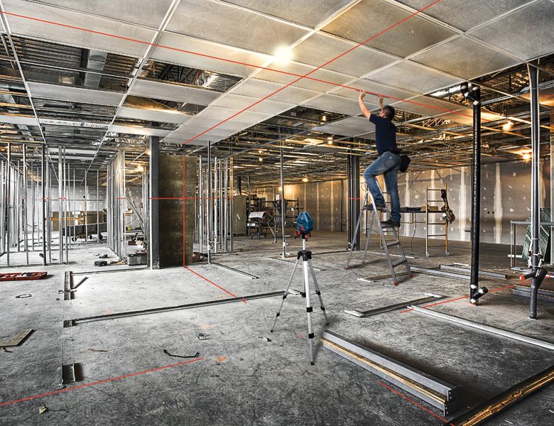 A person on a ladder installs ceiling panels in a large room using a laser leveling tool.