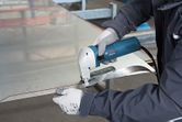 A person wearing safety equipment uses an electric shear to cut a marked metal sheet.