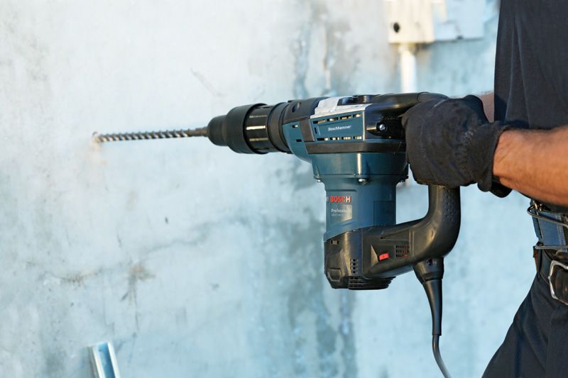 A person wearing safety equipment drills into concrete with a rotary hammer.