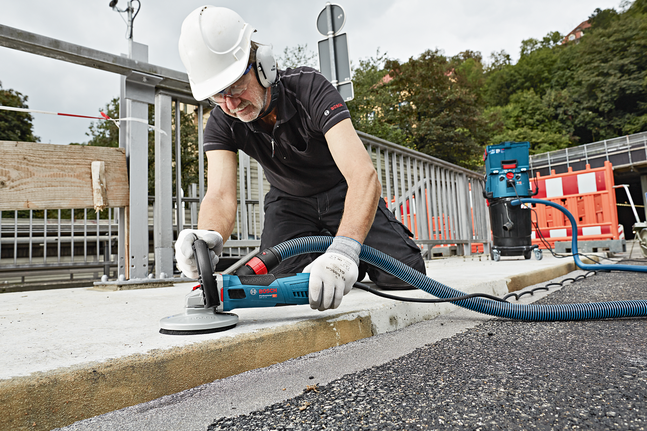 A person wearing safety equipment grinds a concrete curb with a connected dust extractor.