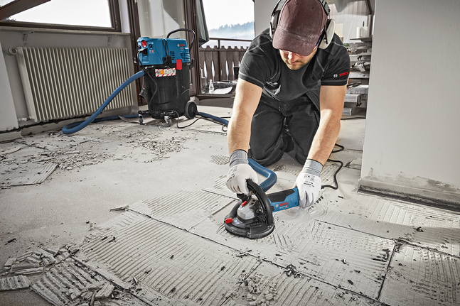 A person wearing safety equipment grinds a concrete floor in a dusty indoor space.