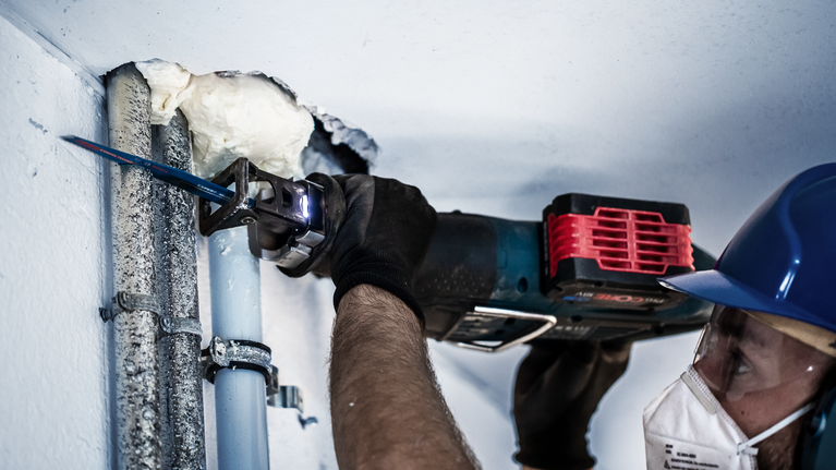 A person wearing safety equipment cuts through metal pipes using a reciprocating saw.