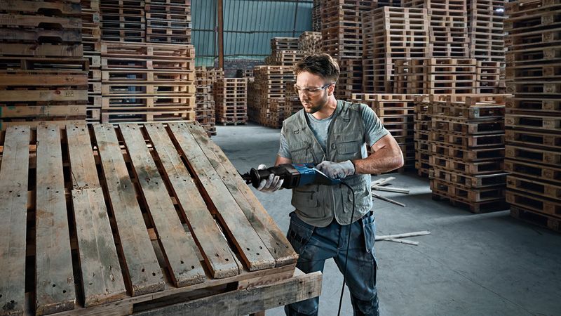 A person wearing safety equipment cuts a wooden pallet using a reciprocating saw.