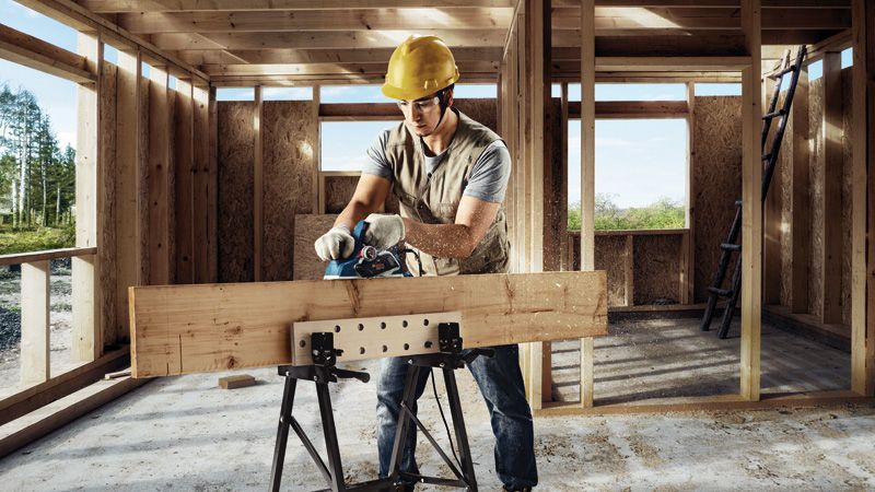 A person wearing safety equipment planes a wooden board inside a building frame.