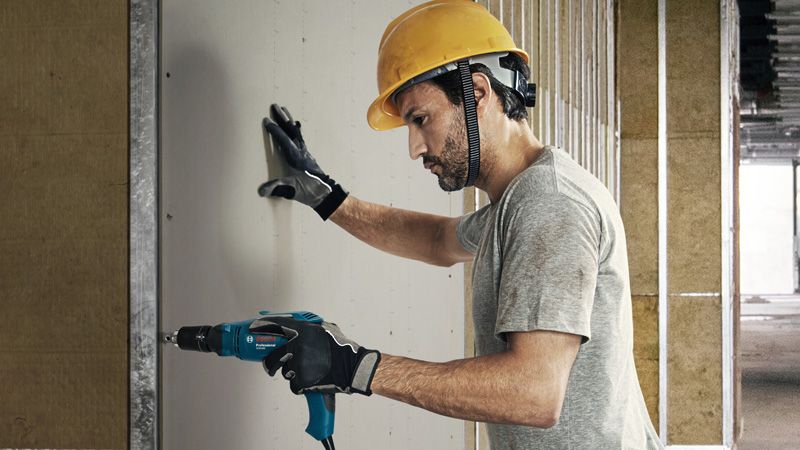 A person wearing safety equipment fastens drywall with a screwdriver.