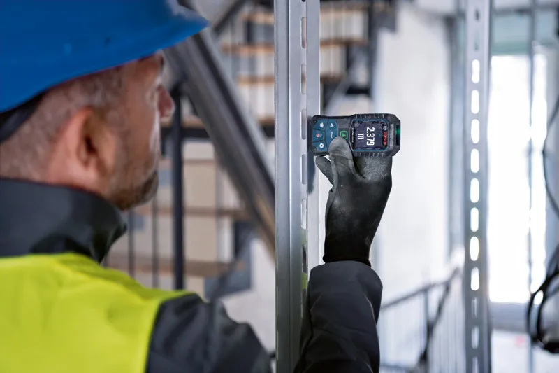 A person wearing safety equipment uses a laser measure to check the distance on a metal frame.