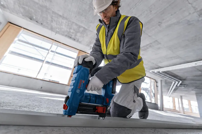 A person wearing safety equipment uses a cordless concrete nailer on a metal track.