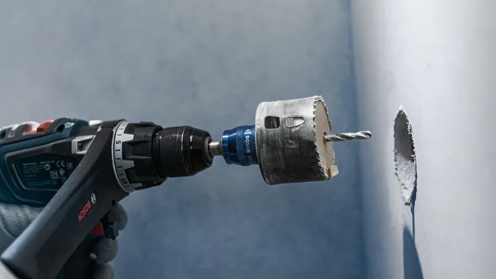 Person wearing safety equipment drills a circular hole into a wall.